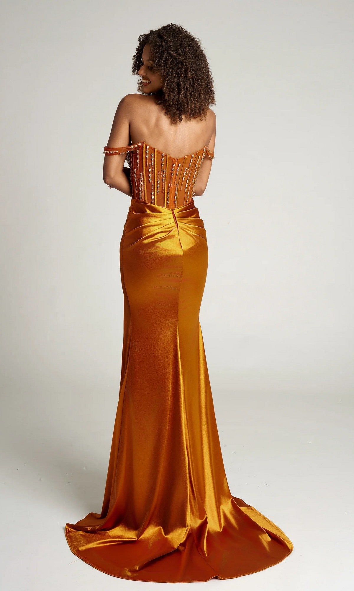 A woman in the Portia and Scarlett PS26046 long formal dress stands facing away, revealing an orange off-the-shoulder design with a fitted beaded corset bodice and a gathered flowing skirt that features a small train.